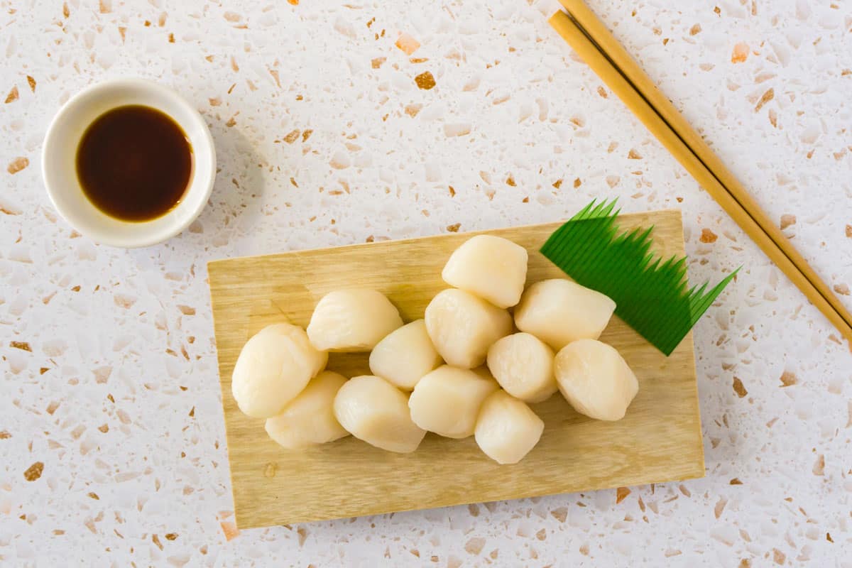 A photo of scallop sashimi on a wooden board with wooden chopsticks and soy sauce.