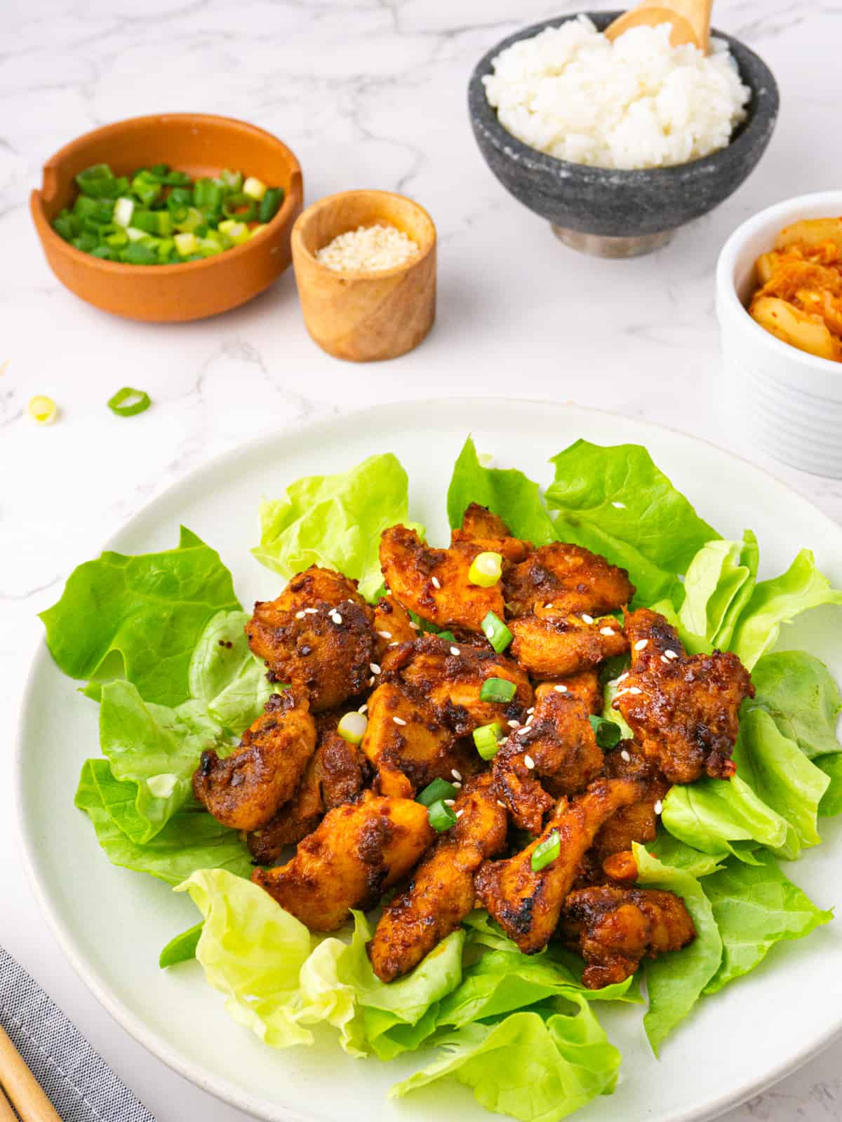 Plate of spicy chicken bulgogi with lettuce wraps and side dishes in the background.