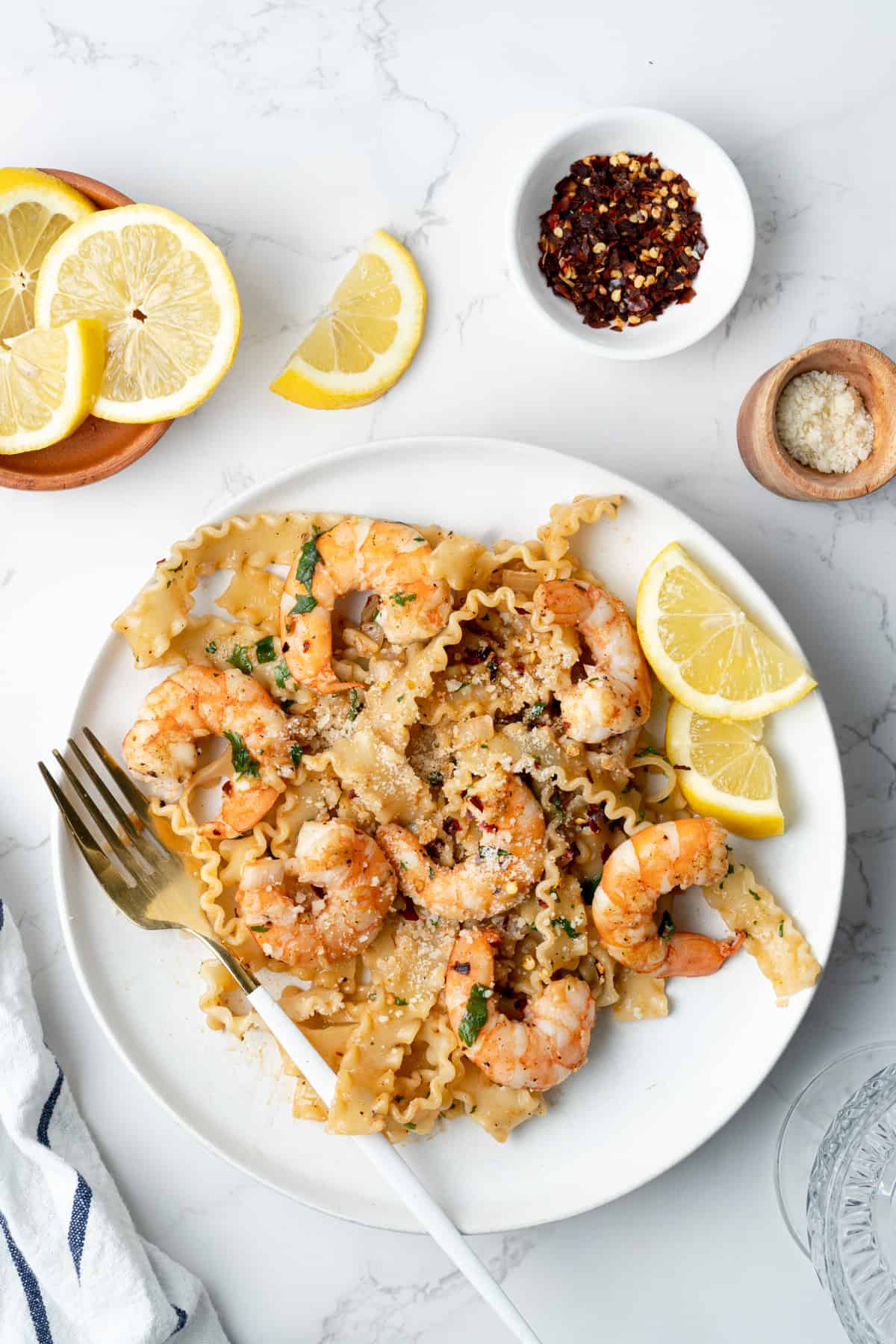 Shrimp scampi pasta in a white plate with lemons, chili flakes, and cheese on the side.